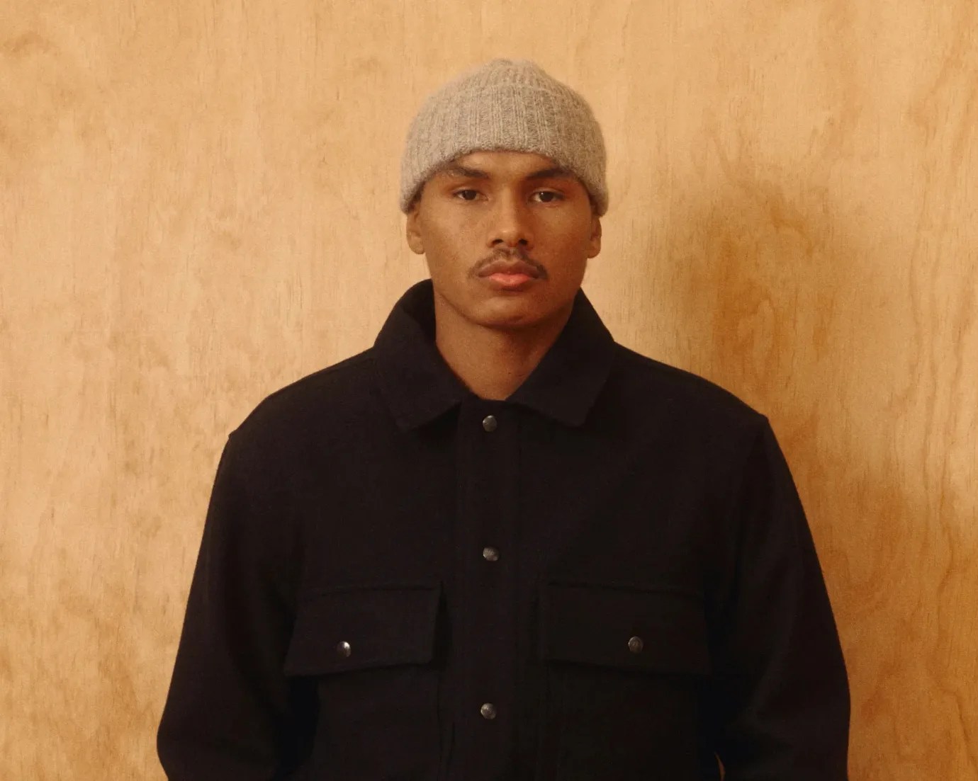 Man wearing a black button-up jacket with flap pockets and a beige knit beanie against a wooden background.