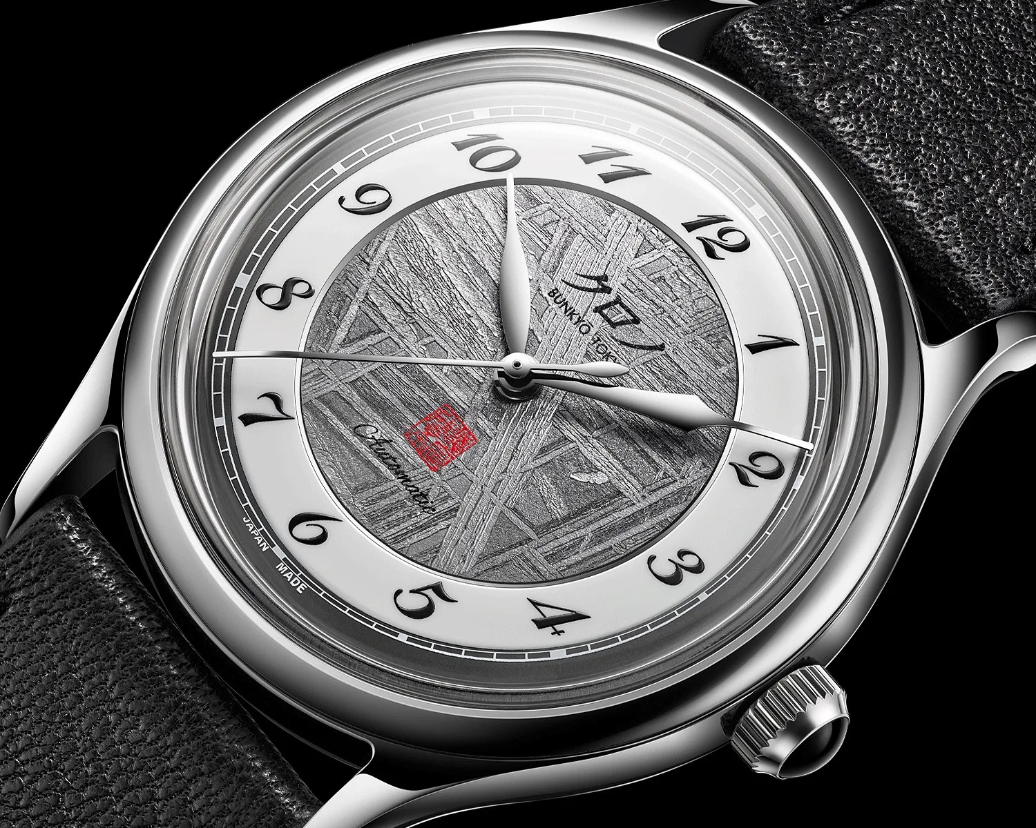 Close-up of a silver wristwatch with a textured gray dial, black Arabic numerals, and a black leather strap.