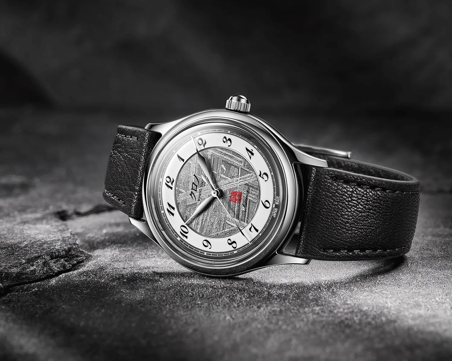 Silver wristwatch with black leather strap and textured gray dial featuring black Arabic numerals and a red square detail.