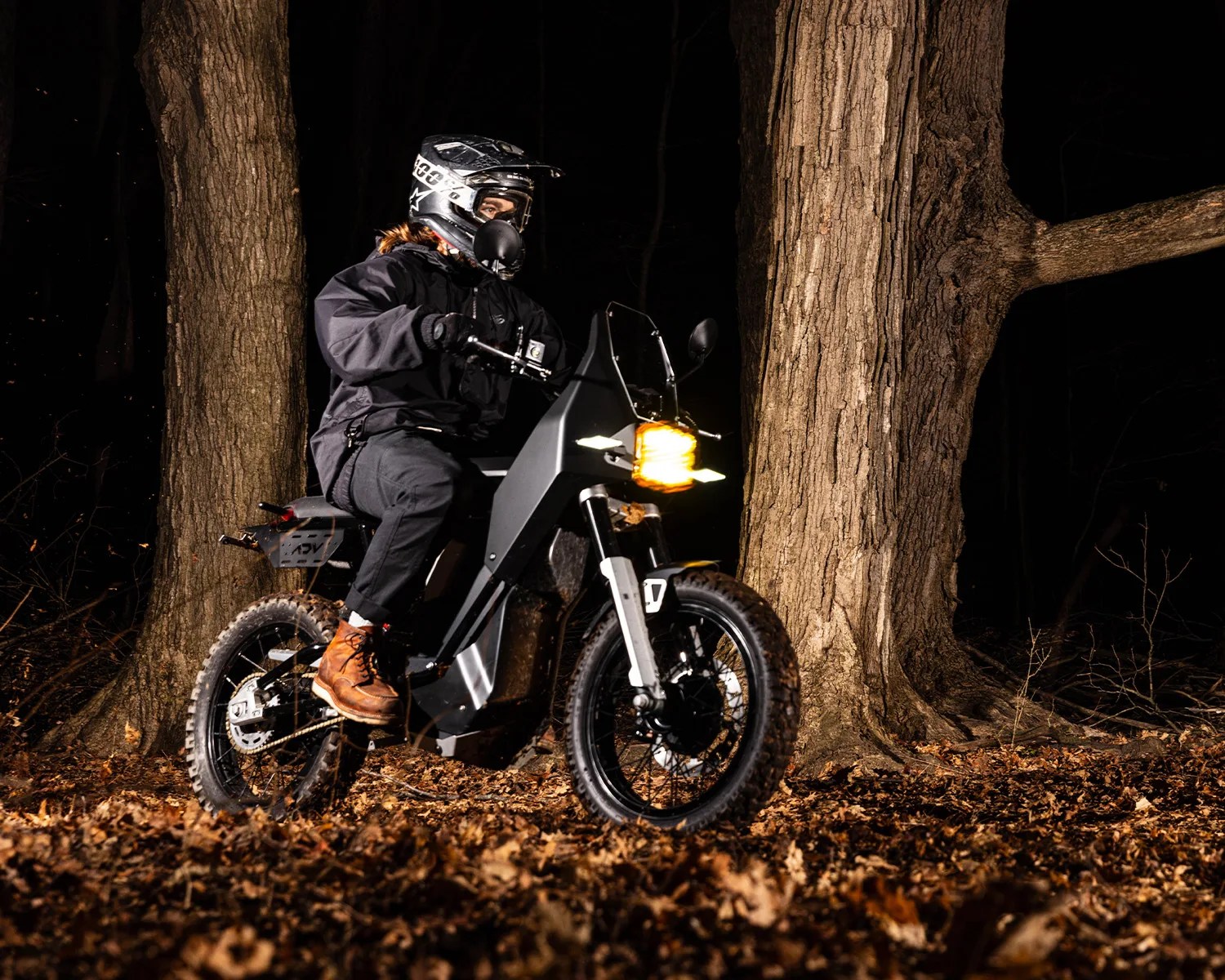Person wearing a black jacket and helmet riding a black off-road motorcycle with a headlight on in a forest at night.