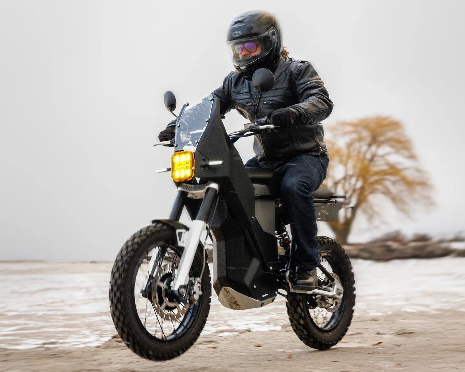 Person wearing a black helmet and leather jacket riding a black electric off-road motorcycle with yellow headlight on a sandy terrain.