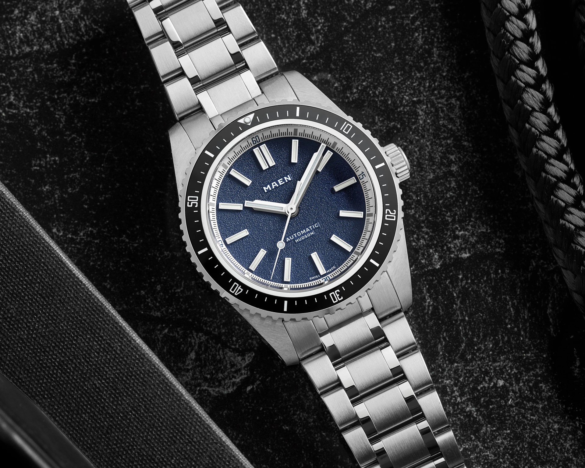 Stainless steel wristwatch with a blue textured dial and black bezel on a dark textured surface.