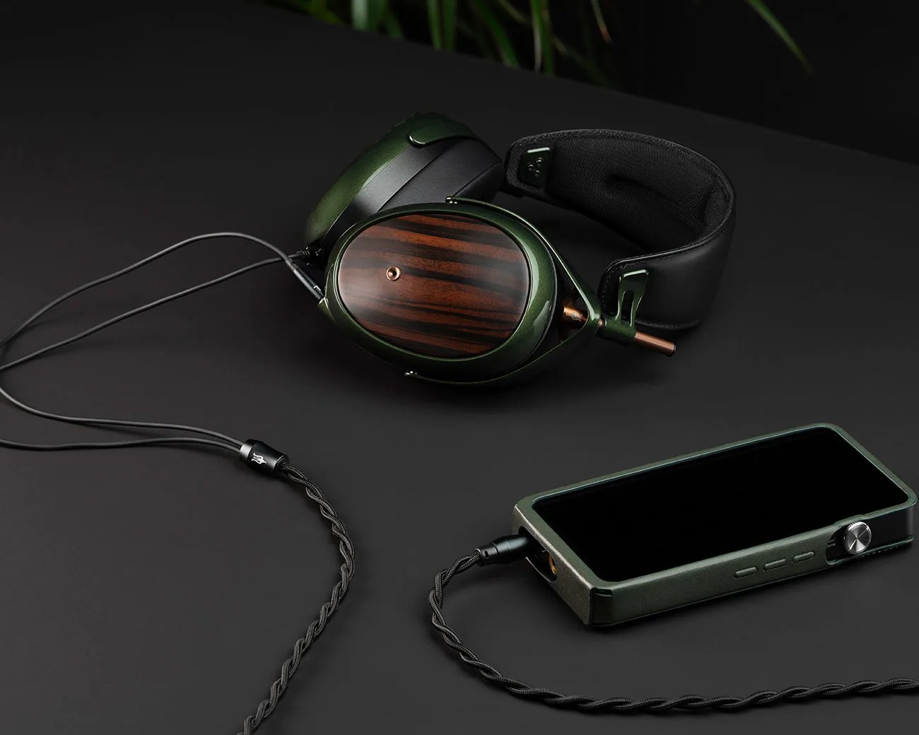 Over-ear headphones with wooden ear cups and green metal frame connected by braided cable to a green digital audio player on a black surface.