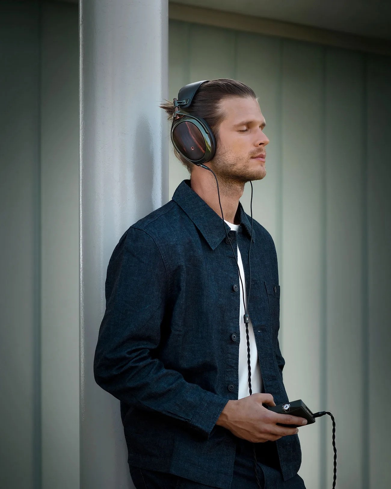 Man wearing dark blue denim jacket and large over-ear headphones, holding a black audio device with eyes closed.