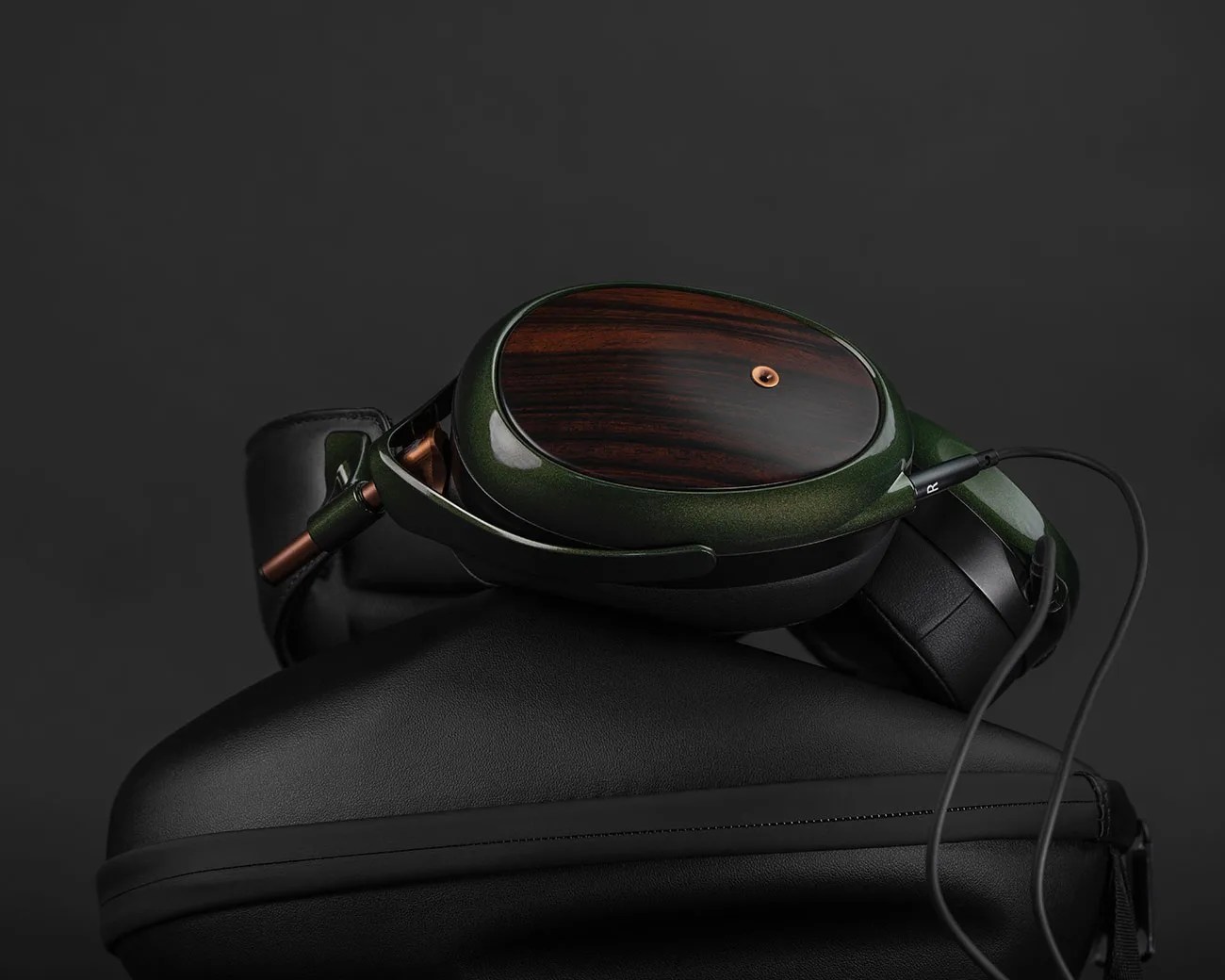 Over-ear headphones with dark green metallic frame, wooden ear cups, and black leather ear pads resting on a black case.