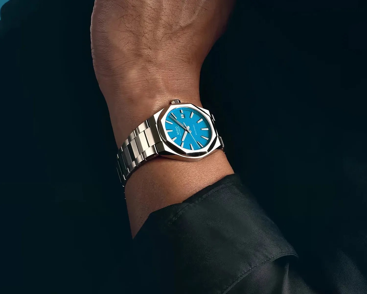 Silver metal wristwatch with a bright blue dial worn on a wrist with a black sleeve.