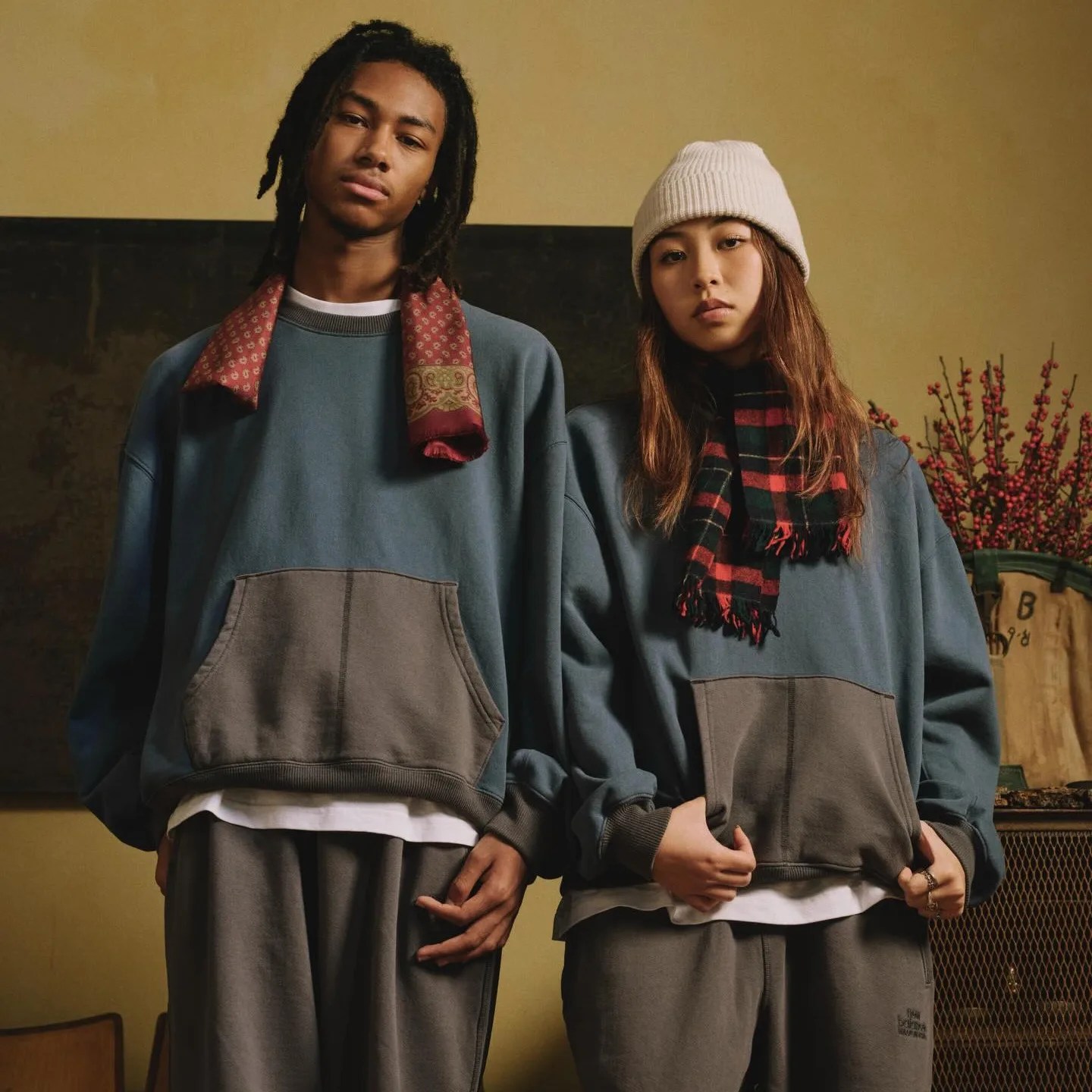 Two people wearing matching oversized blue and gray sweatshirts with front pockets, paired with gray pants, one with a red patterned scarf and the other with a red and black plaid scarf and beige knit beanie.