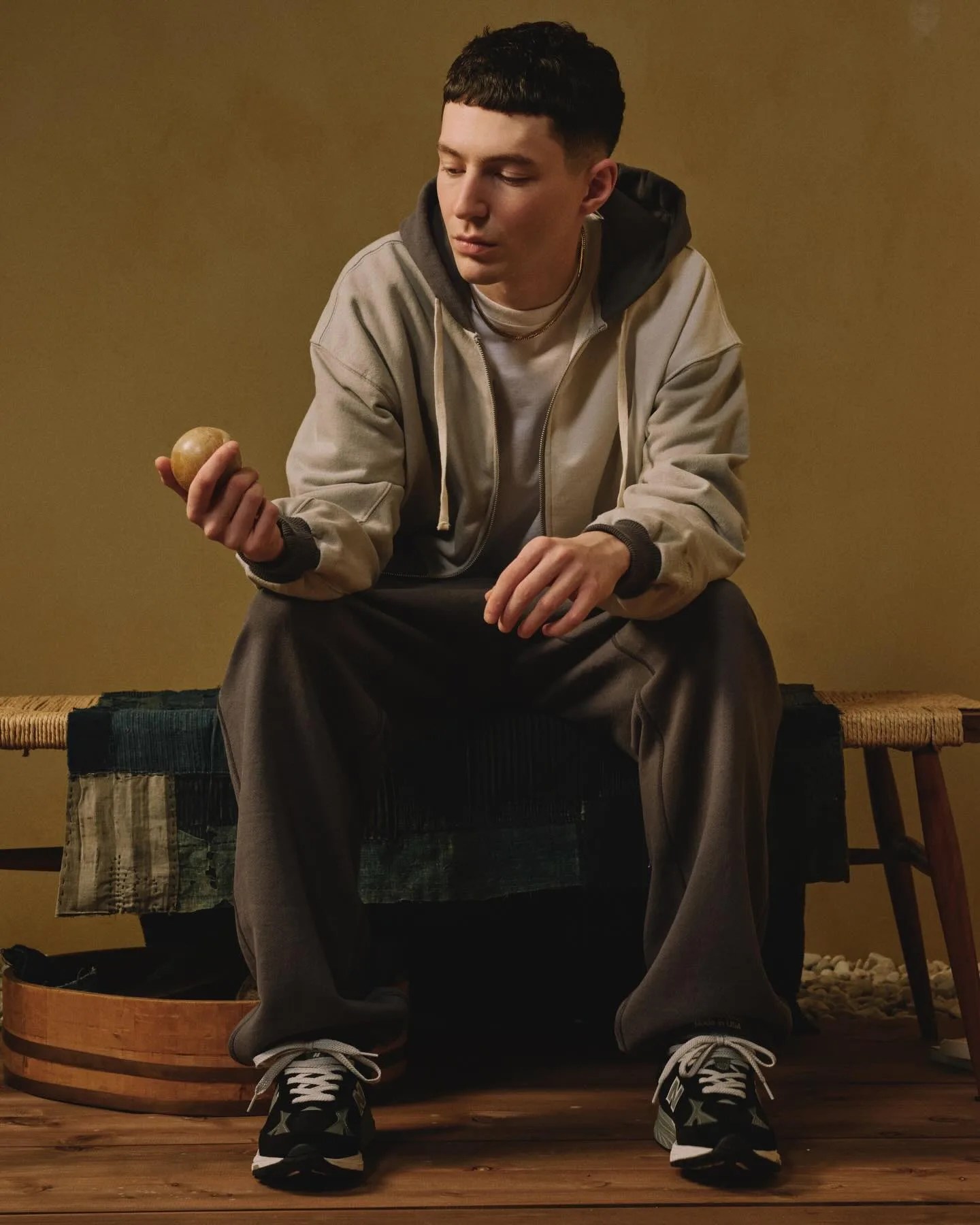 Person wearing a beige and gray hoodie with dark sweatpants and black sneakers sitting on a bench holding a round object.
