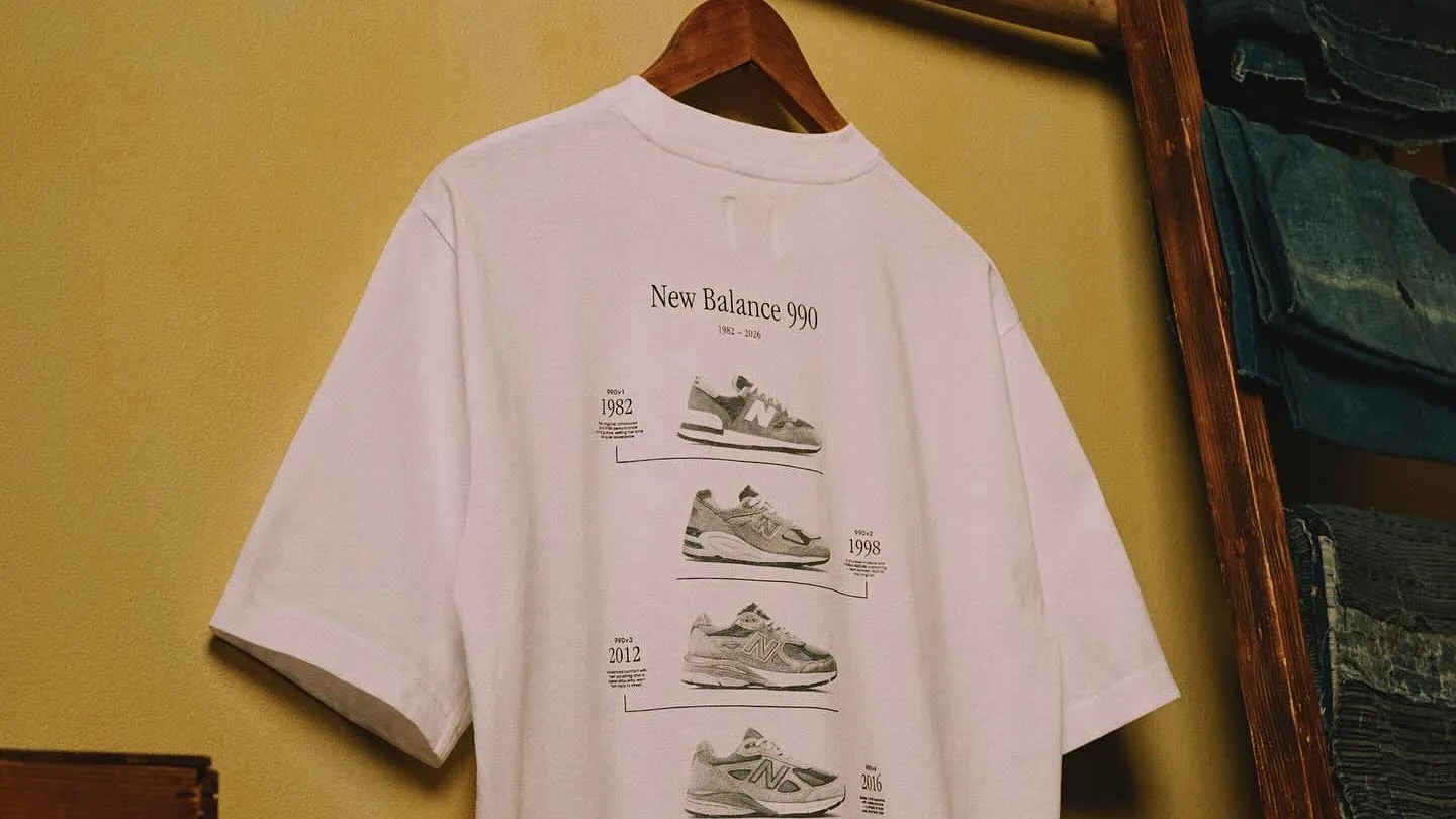 White T-shirt hanging on a wooden hanger with New Balance 990 sneaker designs and years printed on the back.