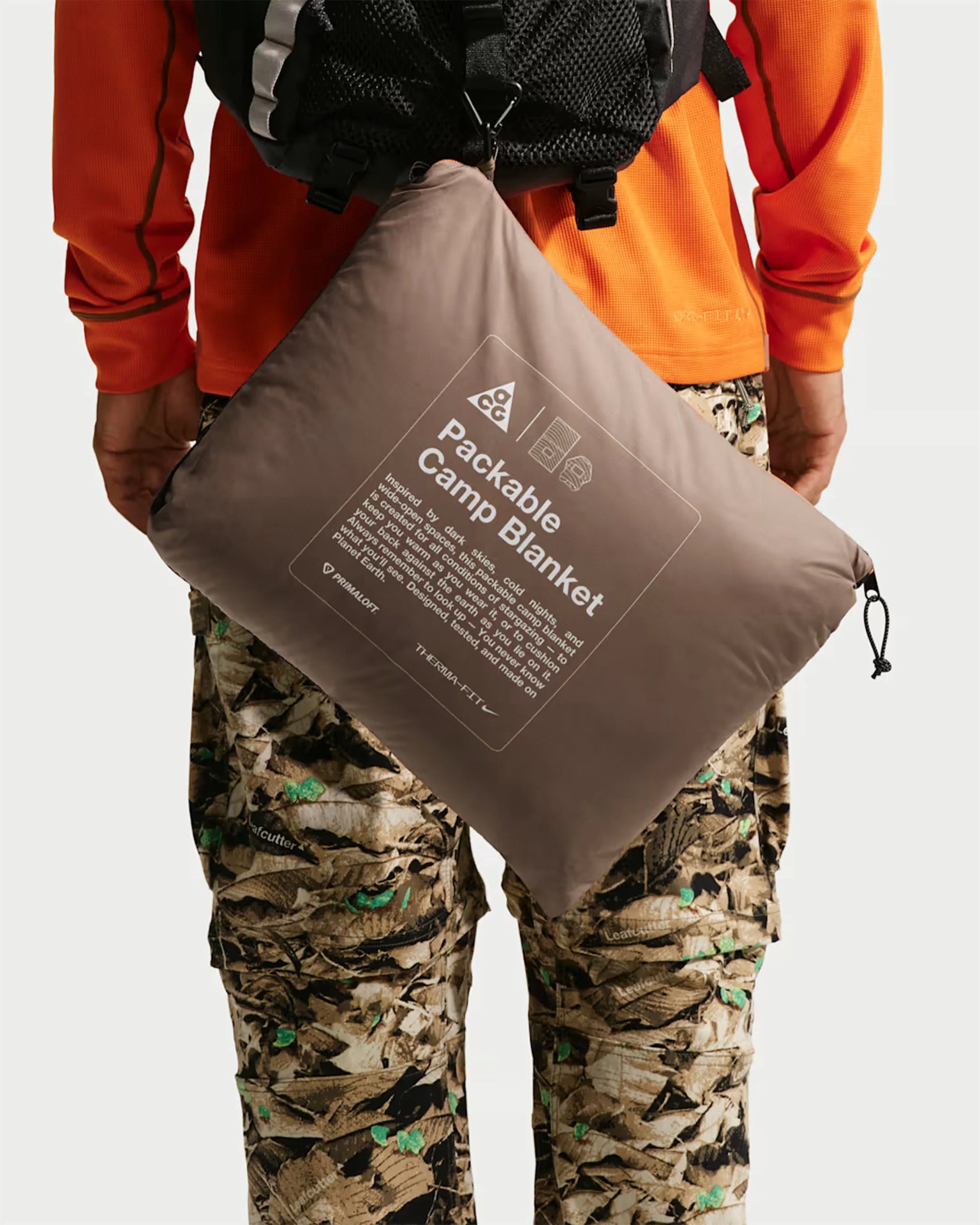 Taupe packable camp blanket in a compact pouch attached to a black backpack worn by a person in an orange top and camo pants.