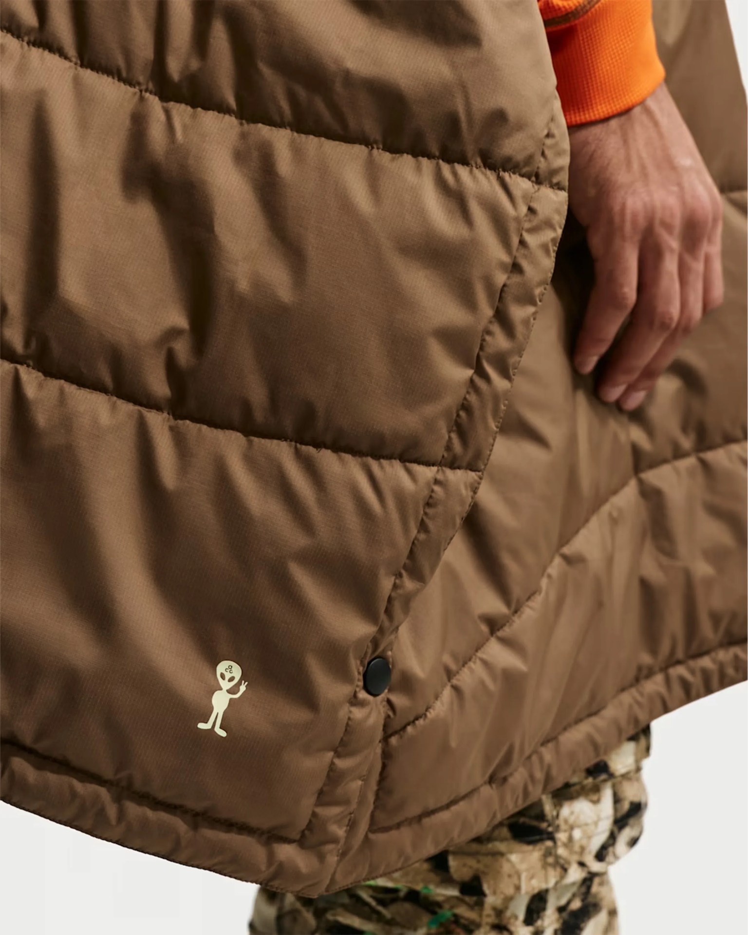 Brown quilted jacket with a small white alien logo near the hem and a black snap button, worn by a person.