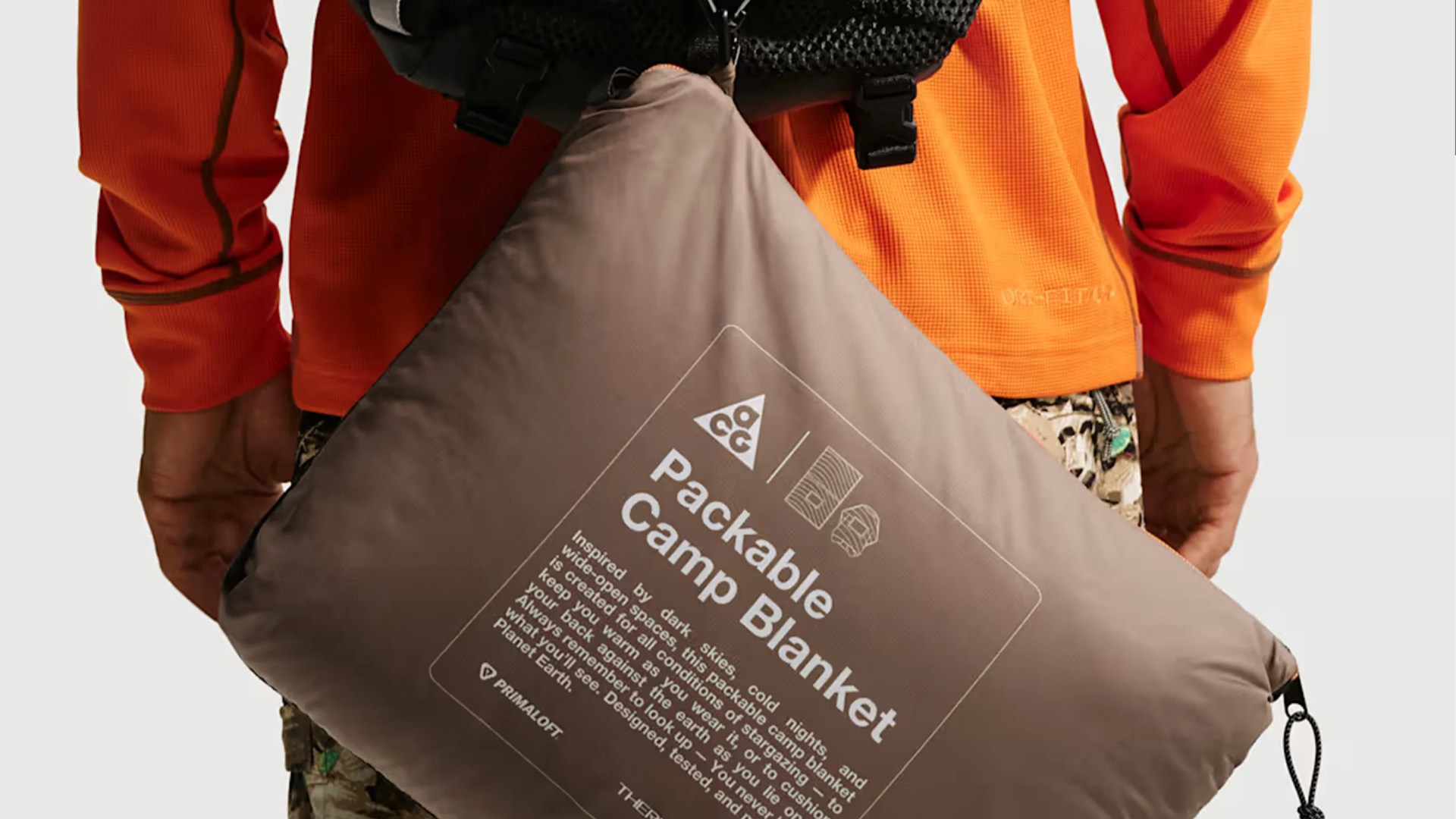 Brown packable camp blanket pouch with white text and logos held by a person wearing an orange jacket and camouflage pants.