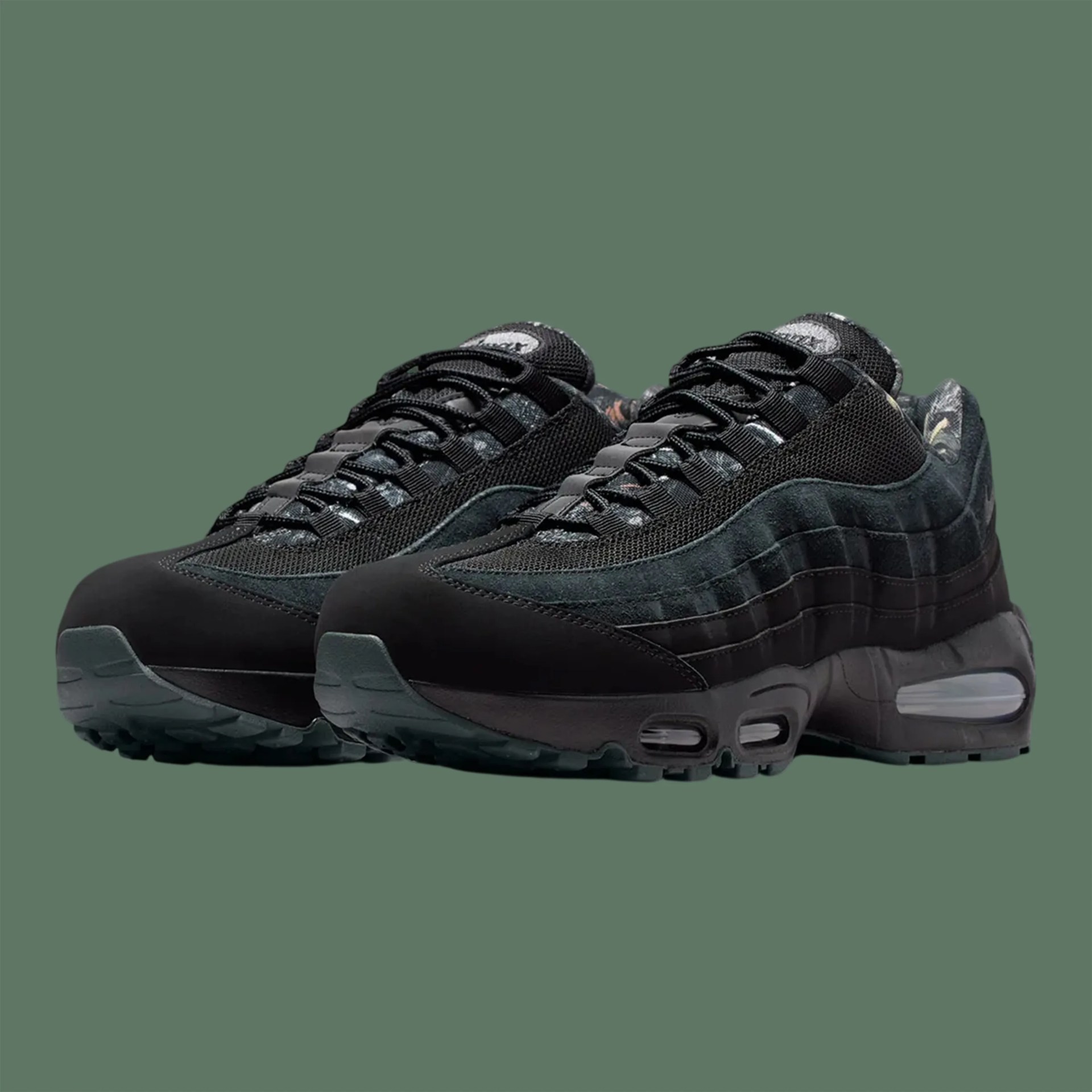 Pair of black and dark grey Nike Air Max 95 sneakers with mesh and suede upper on a green background.