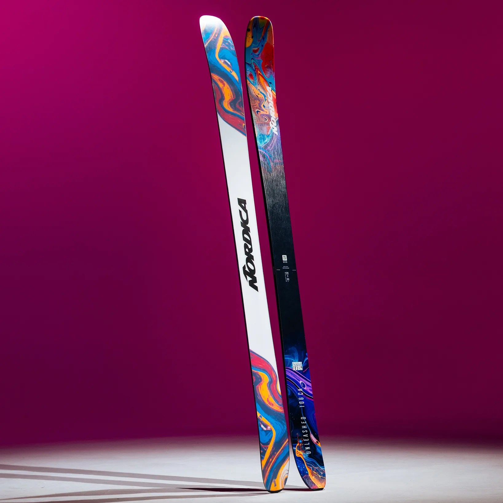Pair of Nordica skis with colorful abstract designs on a white and black background standing upright against a purple backdrop.