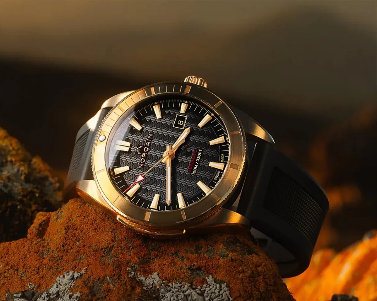 Gold-tone wristwatch with black textured dial, luminous markers, date window, and black rubber strap resting on a rock.