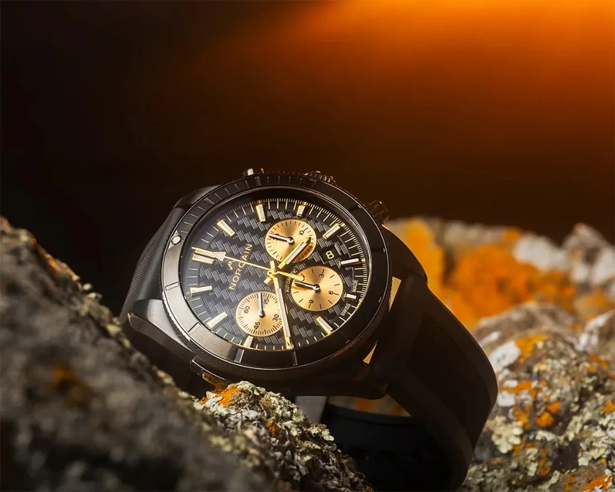 Black Norqain wristwatch with gold chronograph dials and markers resting on rocky surface with orange lichen.