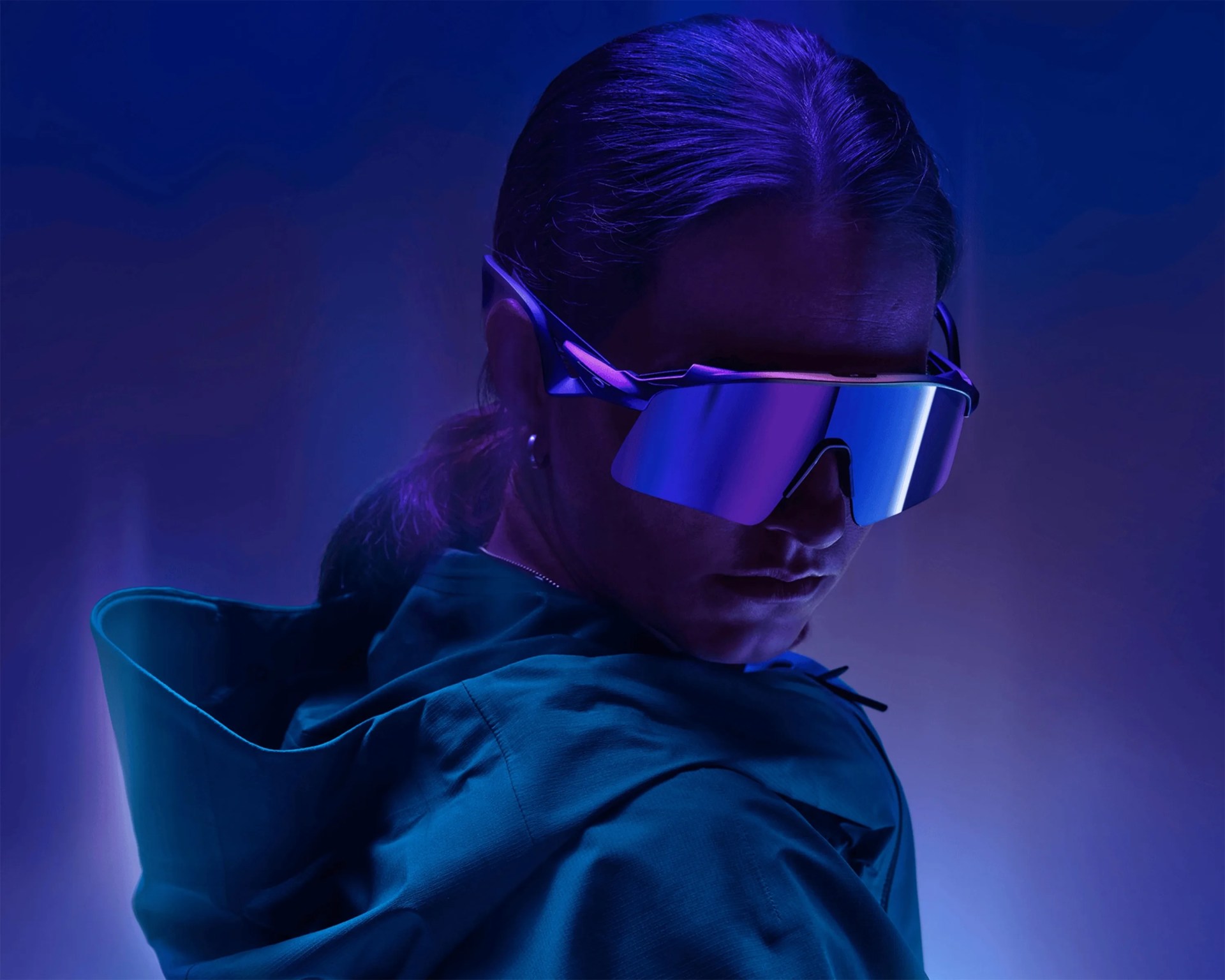 Person wearing large, reflective blue-purple visor sunglasses and a teal jacket with a hood in purple-blue lighting.