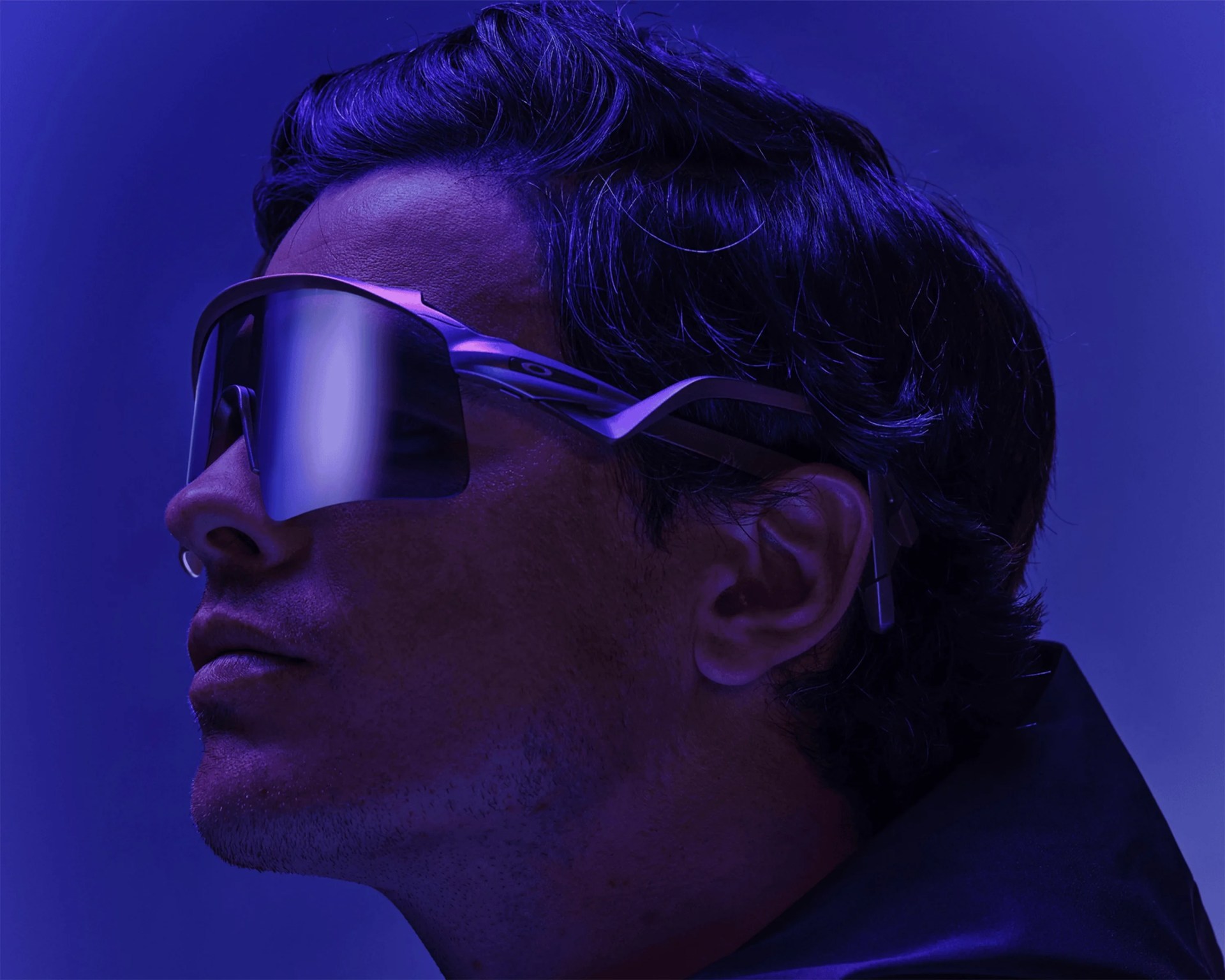 Man wearing large, reflective, wraparound sunglasses with a dark frame in blue lighting.