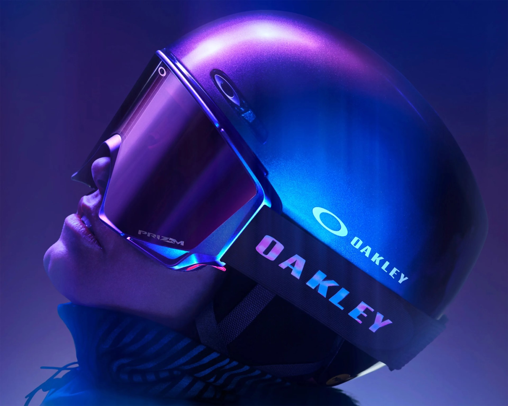 Person wearing a metallic blue Oakley helmet and reflective purple Oakley Prizm ski goggles.
