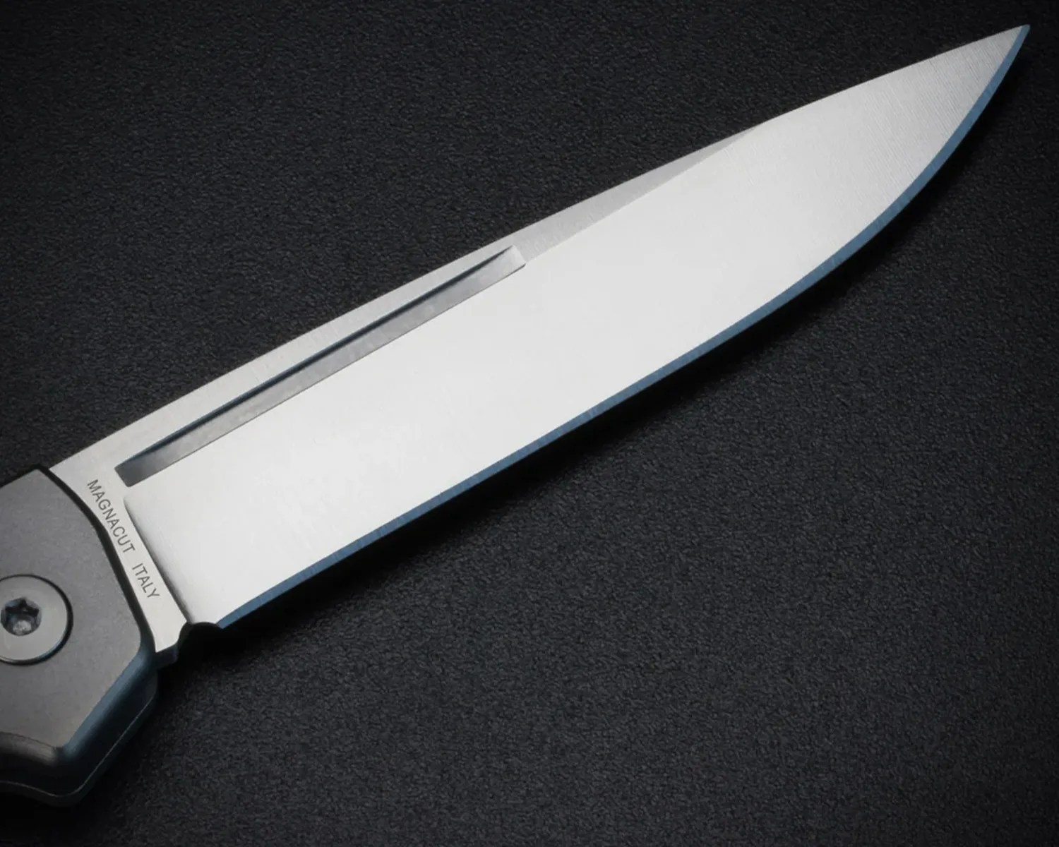 Close-up of a silver Magnacut Italy knife blade with a straight edge on a black textured surface.