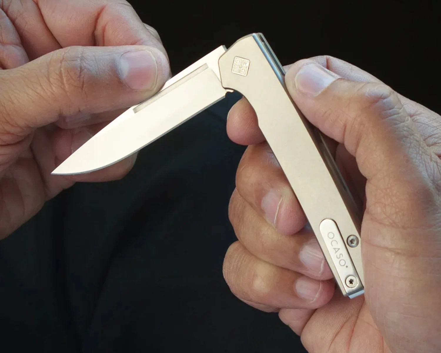 Hand holding a beige folding knife with a partially open blade and "OCASO" engraved on the handle.