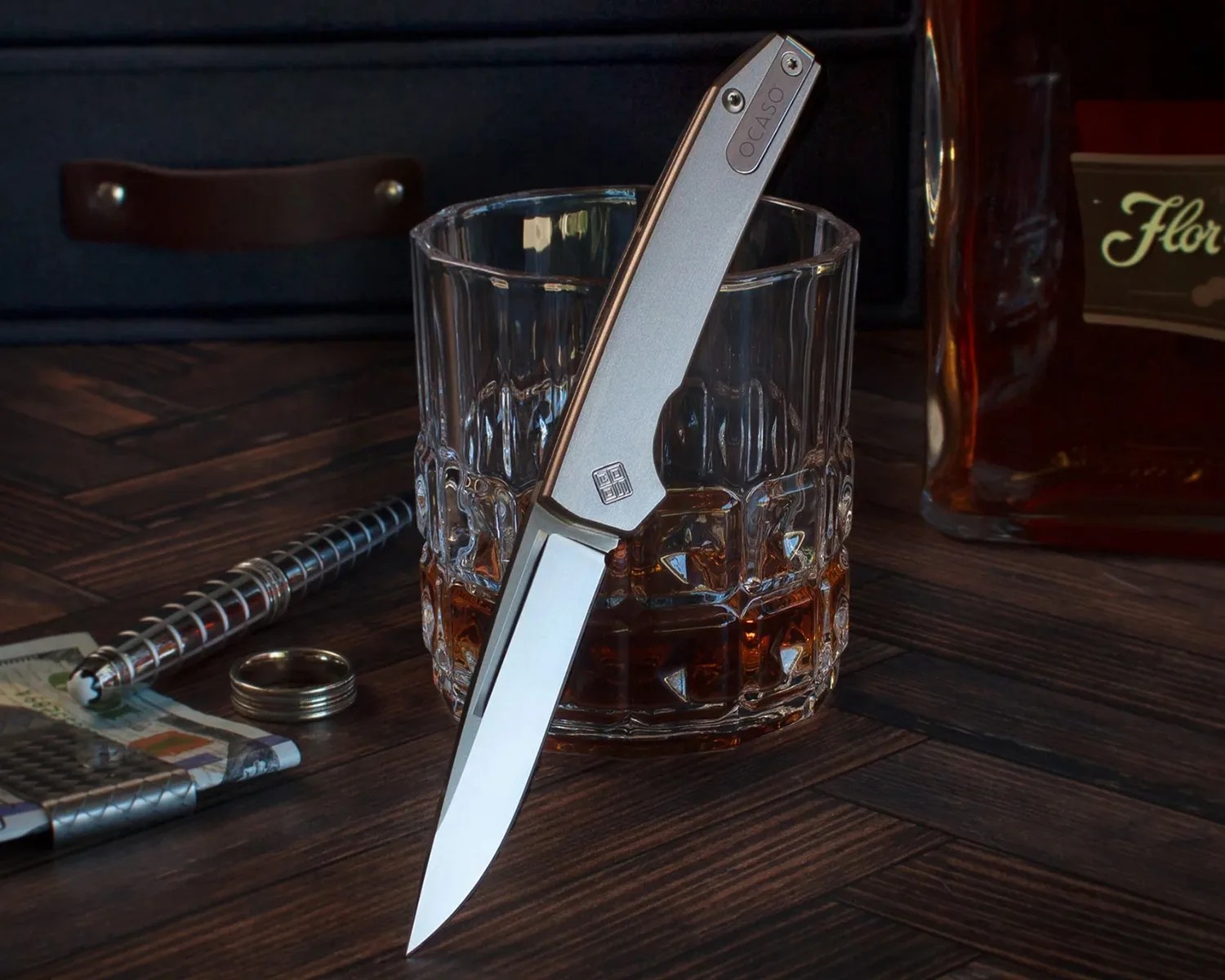 Silver folding knife with a straight blade standing in a glass of amber liquid on a wooden surface.