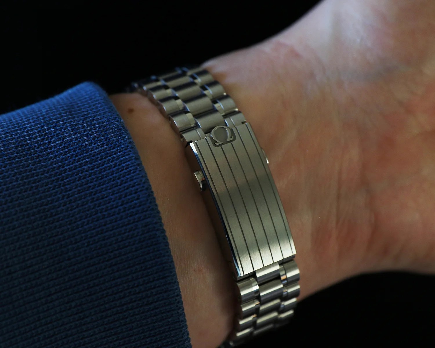 Silver metal watch bracelet with Omega logo clasp worn on a wrist with a blue sleeve.