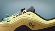 Close-up of a yellow and brown On running shoe with cream laces and a green pull tab against a blue-gray background.