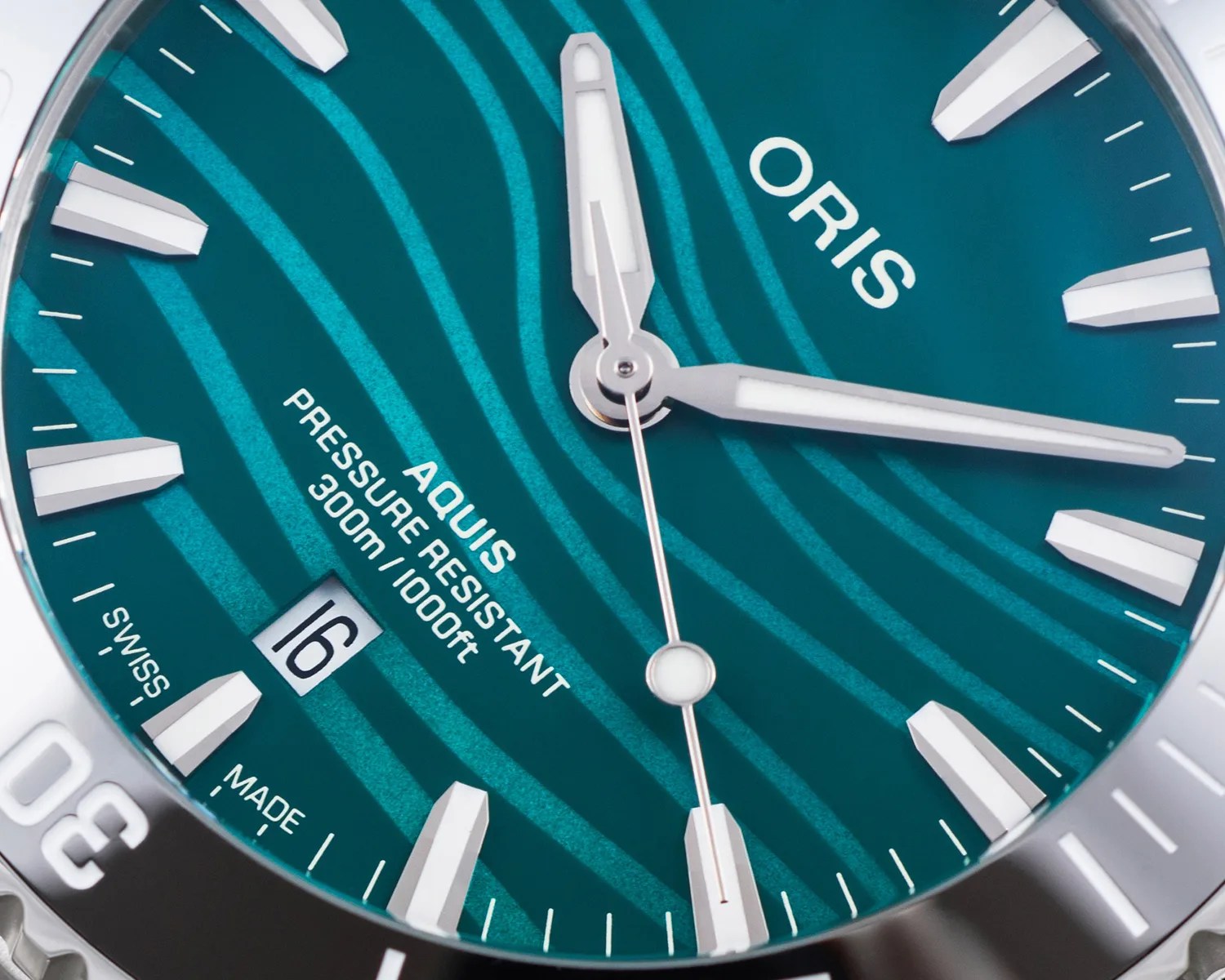 Close-up of an Oris Aquis watch face with teal dial, white hour markers, date window at 6 o'clock, and silver hands.