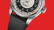 Close-up of a silver automatic wristwatch with a black and white dial, red date hand, and black textured strap on a red background.
