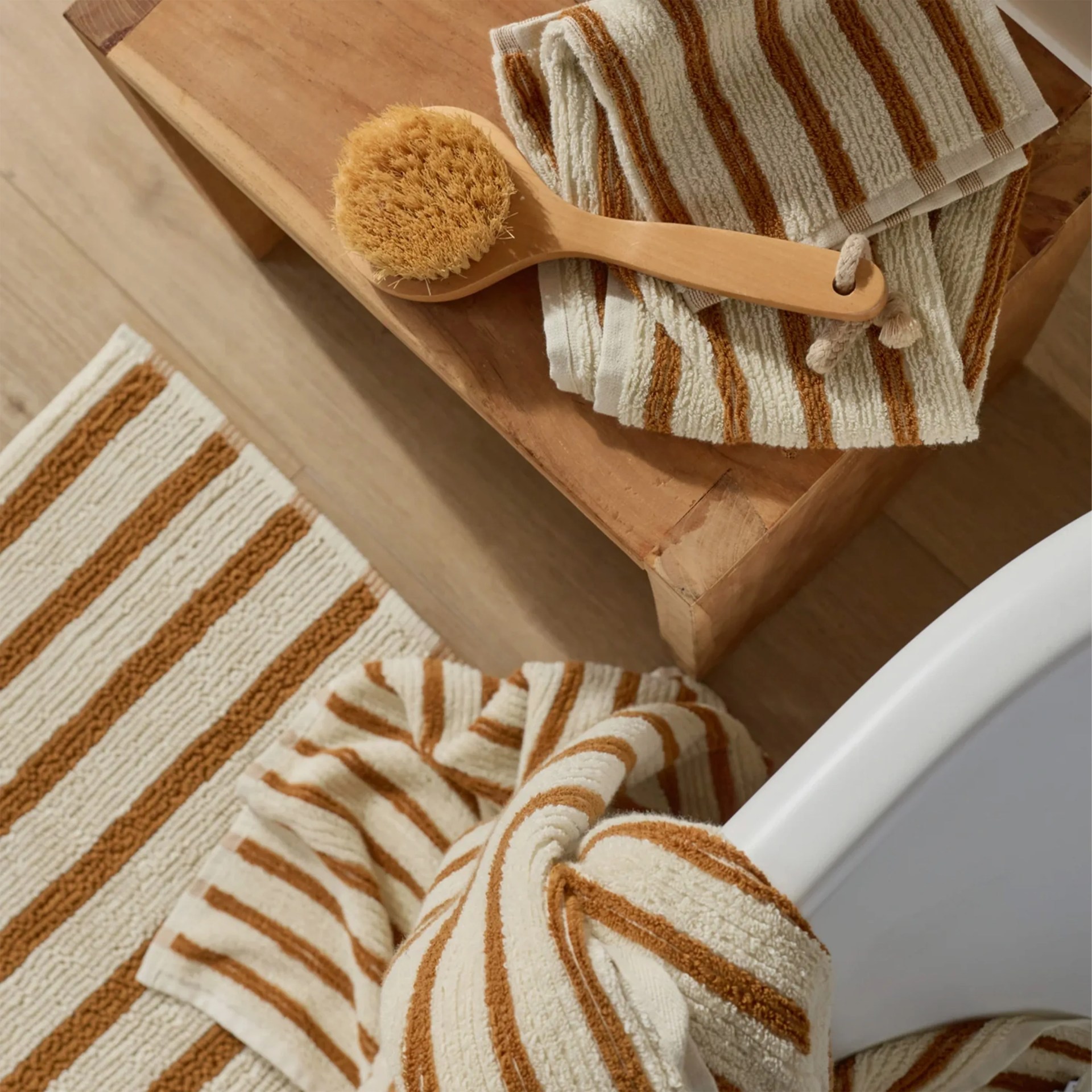 Brown and white striped bath towels and rug with a wooden bath brush on a wooden bench next to a white bathtub.