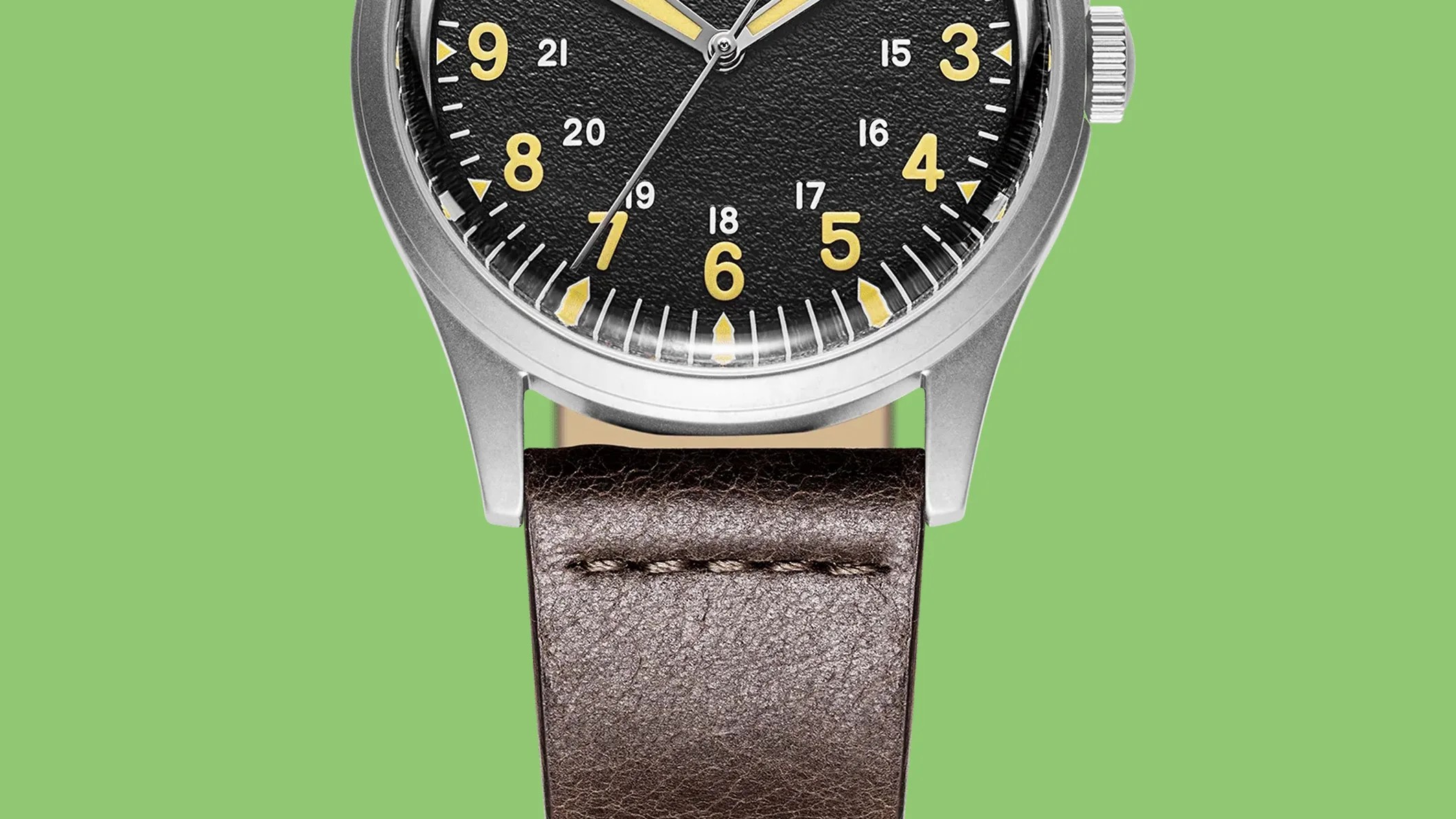 Close-up of a wristwatch with a black textured dial, yellow numerals, silver case, and brown leather strap against a green background.