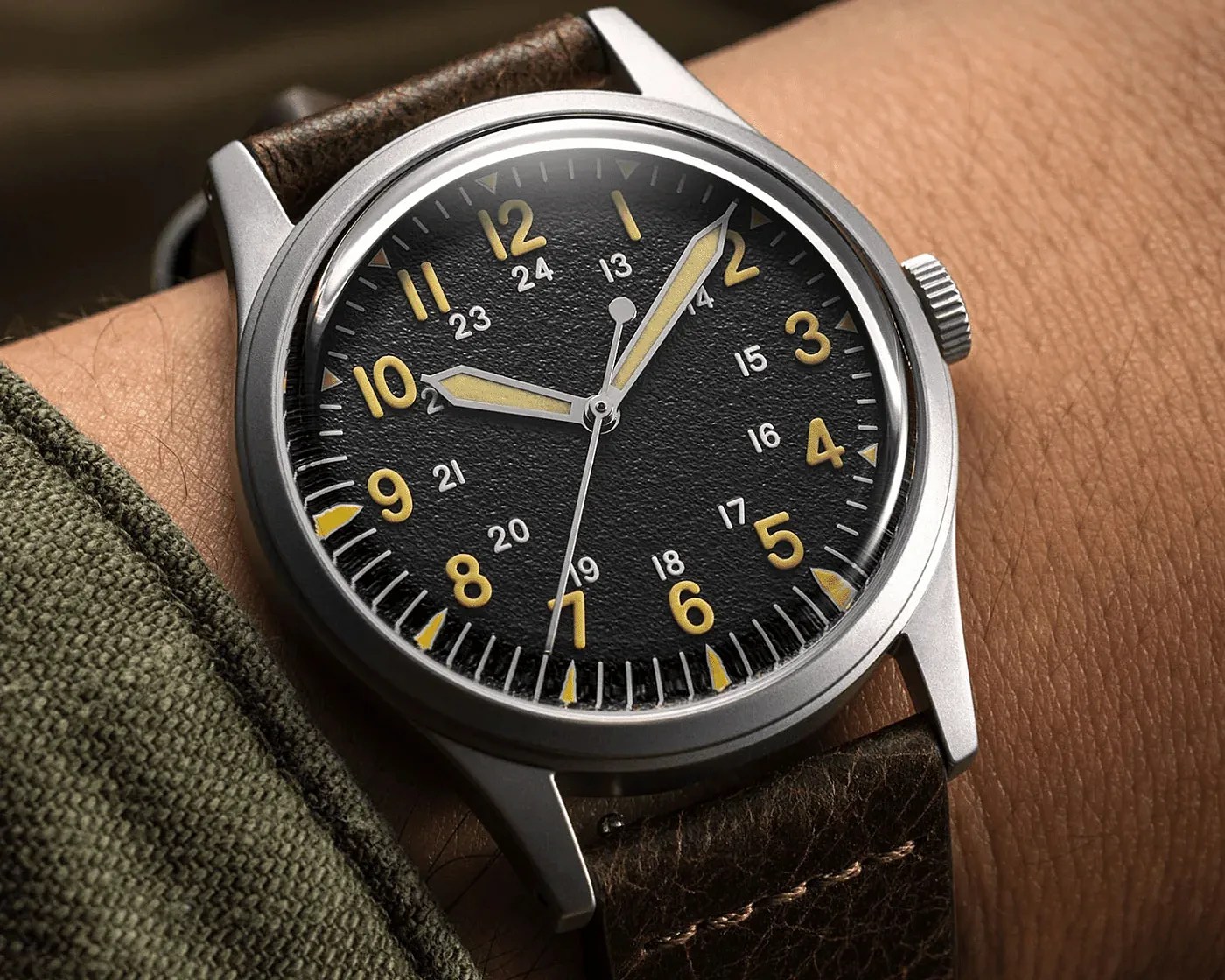 Close-up of a wristwatch with a black textured dial, yellow numerals, silver case, and brown leather strap on a wrist.