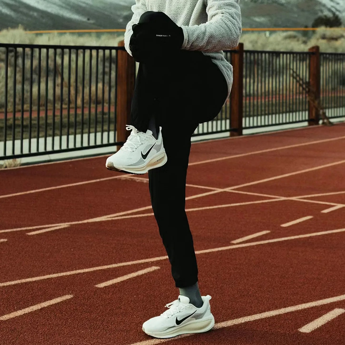 Person in black pants and white Nike sneakers stretching one leg on a red running track.