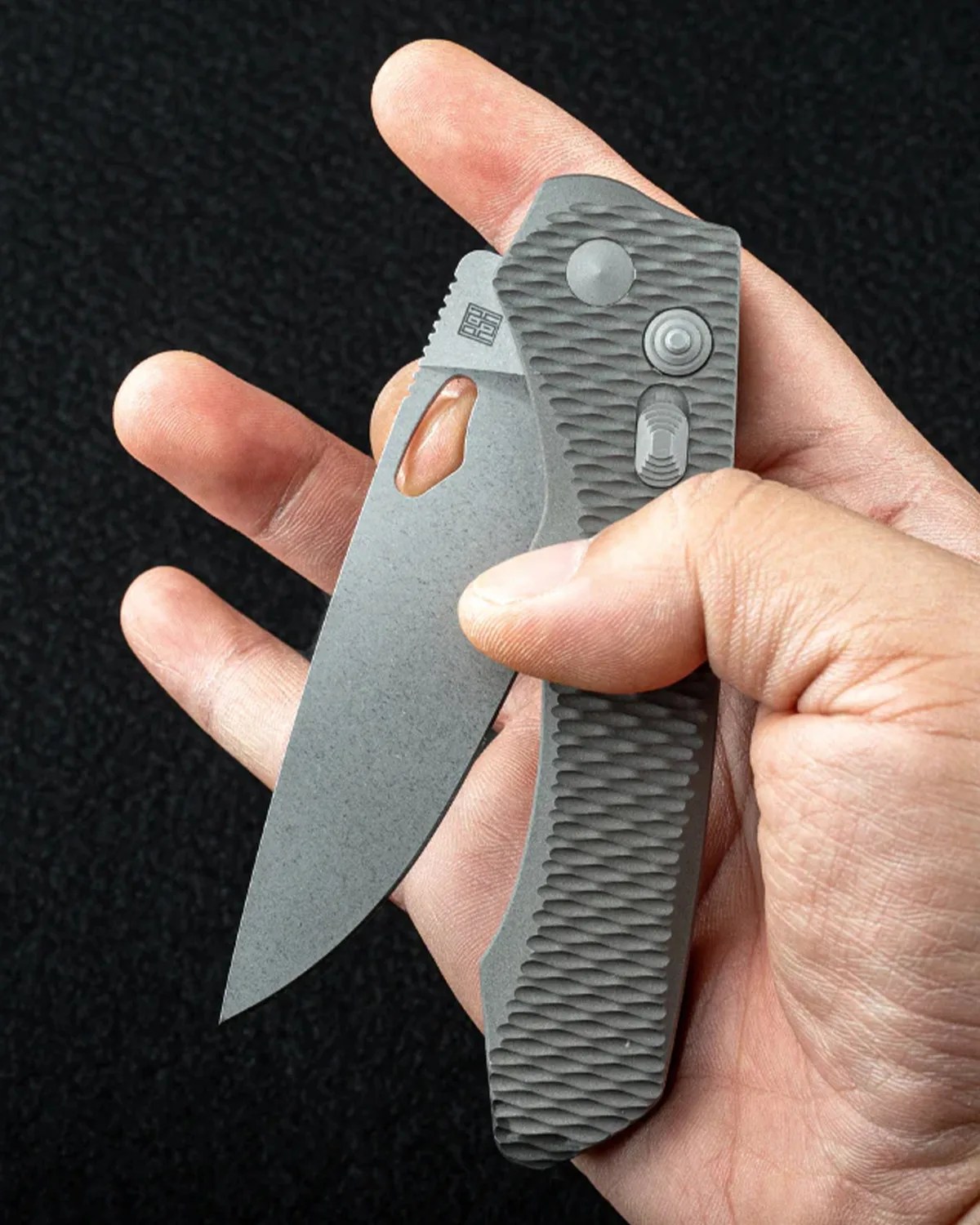 Gray textured folding knife with partially open blade held in a hand against a black background.