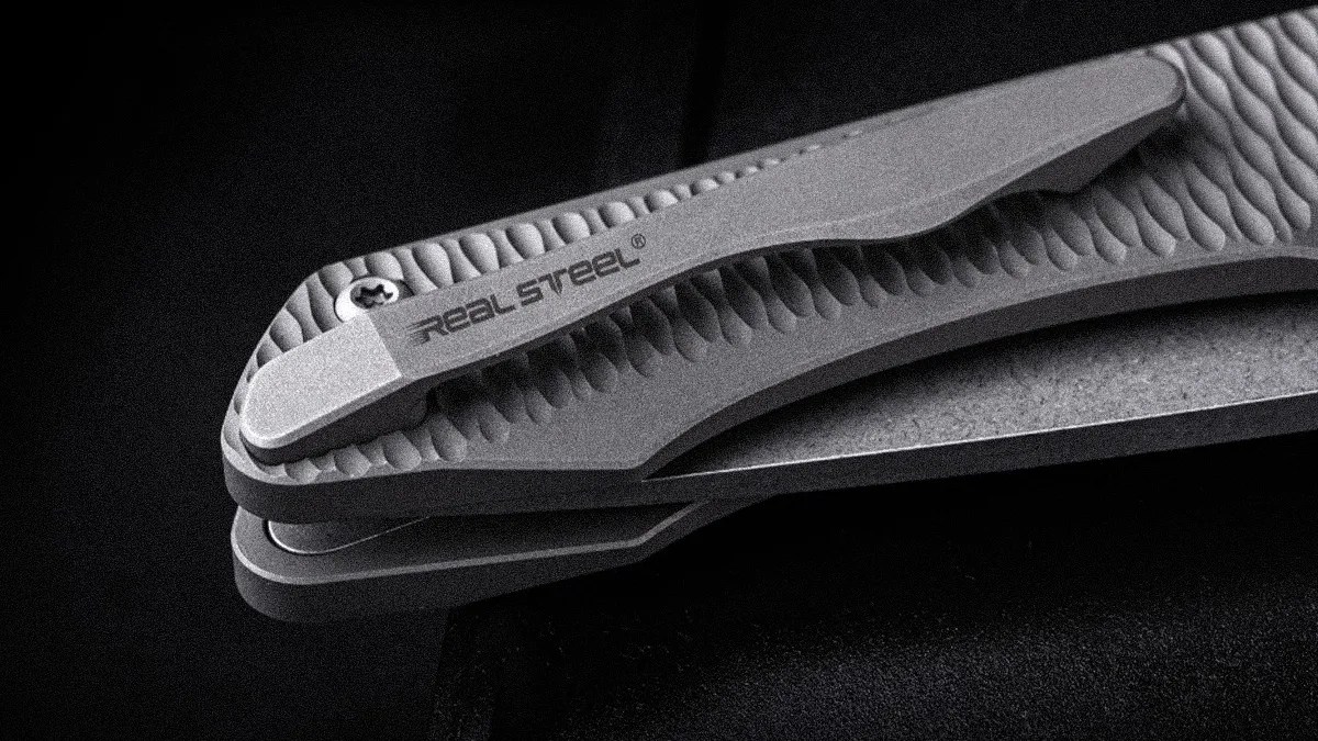Close-up of a textured gray folding knife handle with a clip labeled "Real Steel" on a black surface.