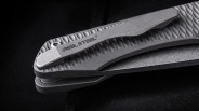 Close-up of a textured gray folding knife handle with a clip labeled "Real Steel" on a black surface.