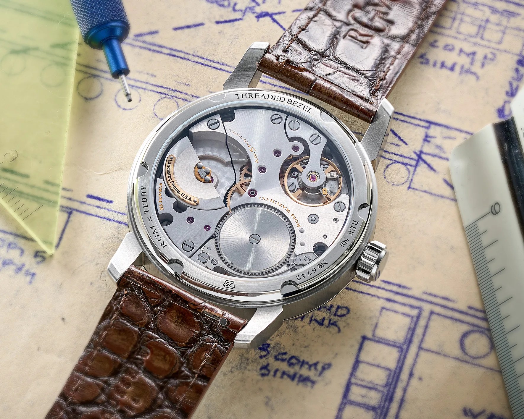 Close-up of a mechanical watch movement with visible gears and screws, housed in a stainless steel case with a brown leather strap, placed on a blueprint with a blue tool and a ruler nearby.
