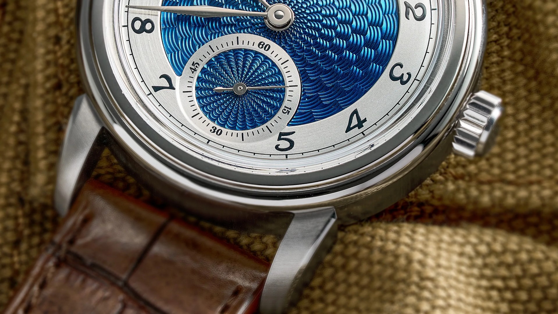 Close-up of a wristwatch with a blue textured dial, silver case, and brown leather strap on a textured fabric surface.