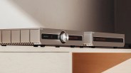 Two silver audio components with black and orange displays, one with a large central knob, placed on a light wooden shelf.