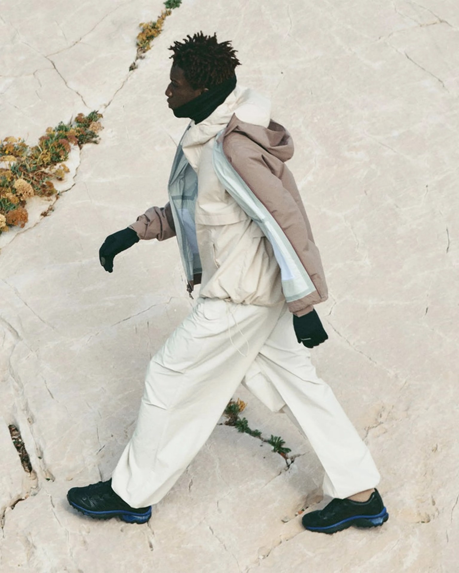 Person walking on a light stone surface wearing beige wide pants, layered beige and brown jackets, black gloves, and black sneakers with blue soles.