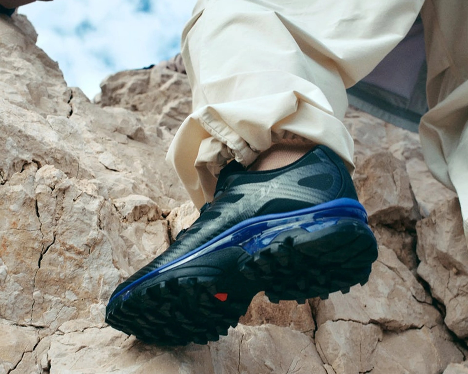 Black and blue trail running shoe with rugged sole worn by a person standing on rocky terrain.