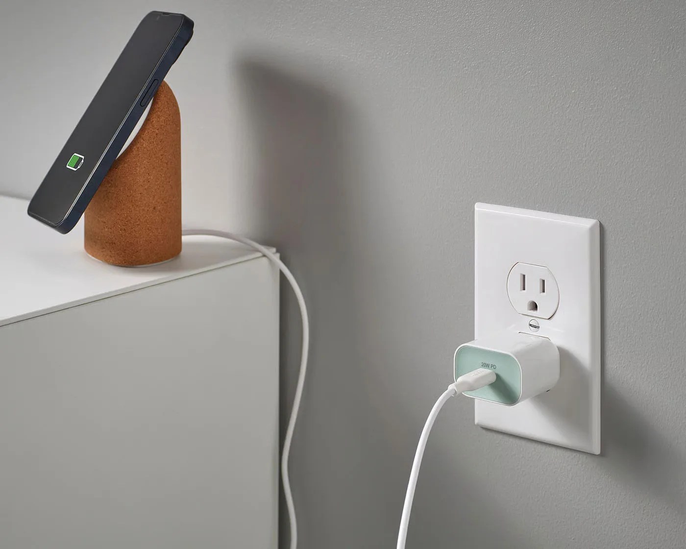 Black smartphone wirelessly charging on a cork stand next to a white wall outlet with a USB-C charger plugged in.
