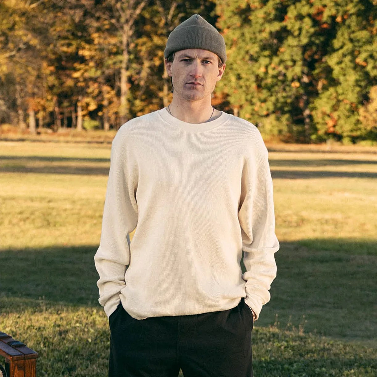 Man wearing a beige knit sweater and olive green beanie standing outdoors with hands in black pants pockets.
