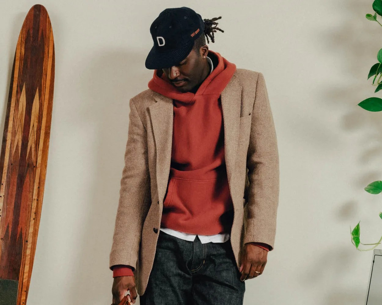 Man wearing a tan blazer over a red hoodie, dark jeans, and a black cap with a white "D" on it.
