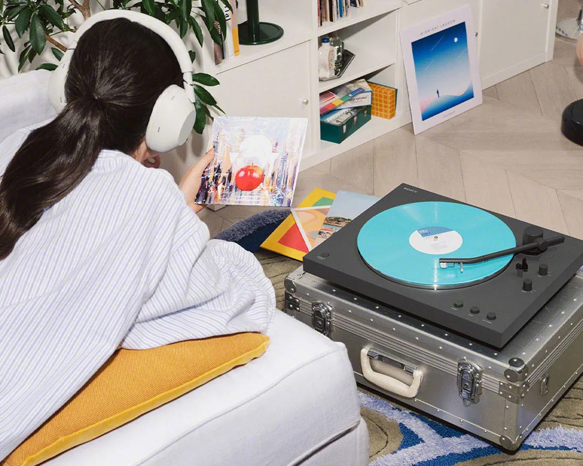 Person wearing white over-ear headphones holding a vinyl record while lying on a white couch with a yellow cushion near a black turntable playing a blue vinyl record.