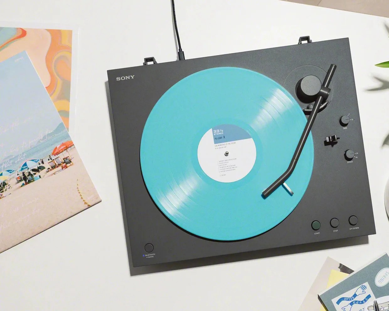 Black Sony turntable playing a turquoise vinyl record on a white surface with album covers nearby.