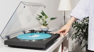 Black turntable with a blue vinyl record and transparent dust cover on a white table, with a hand pressing a button.