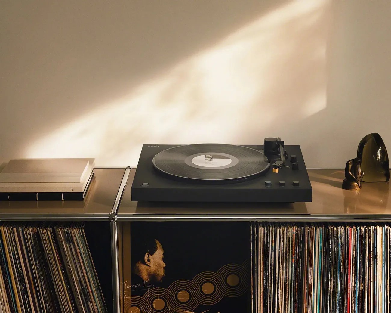Black Sony turntable playing a vinyl record on a metal shelf with stacked books and vinyl records below.