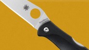 Close-up of a Spyderco folding knife blade with a black handle on a mustard yellow background.